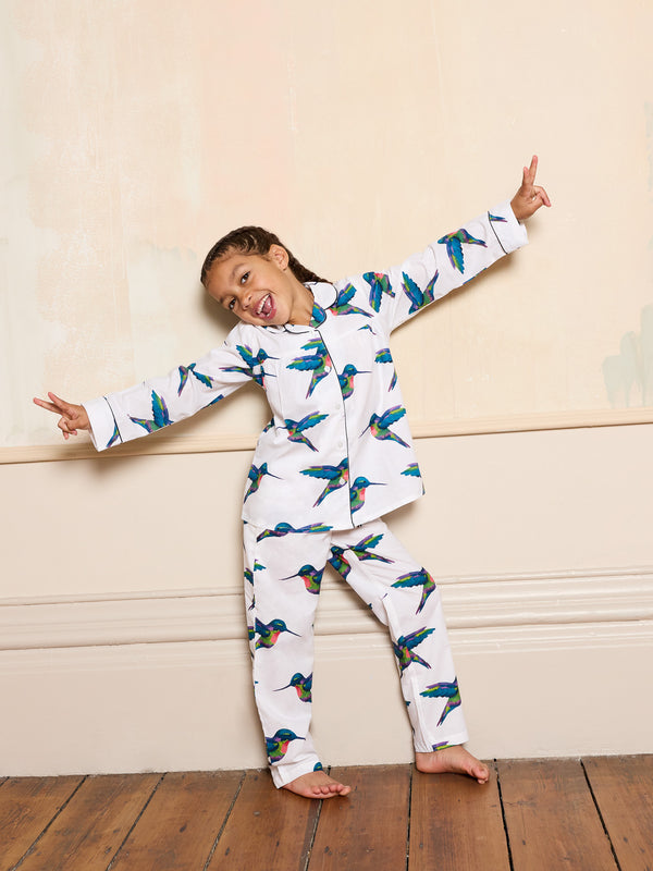 Child wearing a white pyjama set with hummingbird print against a beige wall.