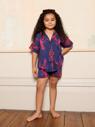 Young girl wearing a navy blue and pink pajama set with lobster pattern against a light wooden panel background.