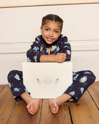 Child sitting on a wooden floor holding a white gift box, wearing a dark blue pyjama set with penguin Christmas themed designs.