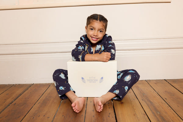 Child sitting on a wooden floor holding a white gift box, wearing a dark blue pyjama set with penguin Christmas themed designs.