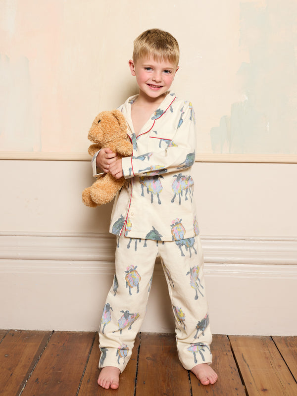 Child wearing a sheep patterned pajama set holding a teddy bear against a light-coloured wall.
