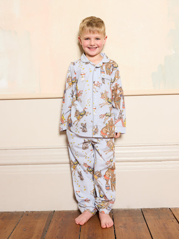 Child wearing a blue circus patterned pajama set standing on a wooden floor with a light-coloured wall background.