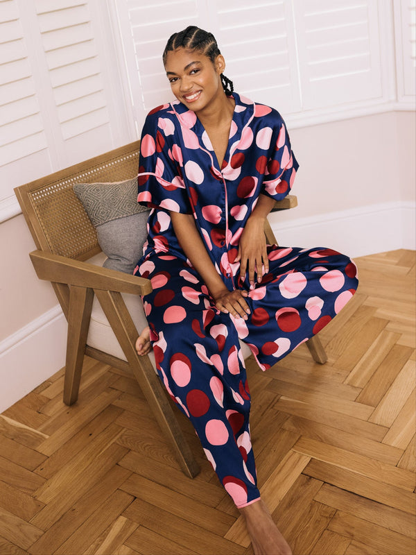 Woman in a polka dot satin pyjama set sitting on a wooden chair indoors.