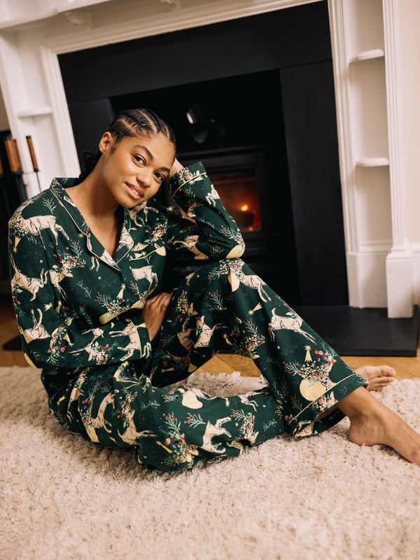 Woman wearing green festive pyjamas sitting on a carpeted floor in front of a fireplace.