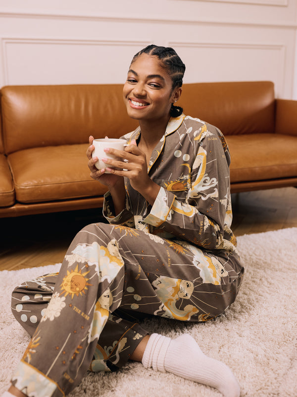 Woman in khaki coloured celestial patterned pyjama set sitting on the floor holding a mug in a living room.