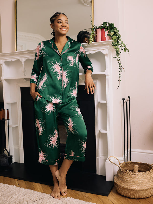 Woman wearing green satin capri pyjamas with pink feather patterns in a cozy room.