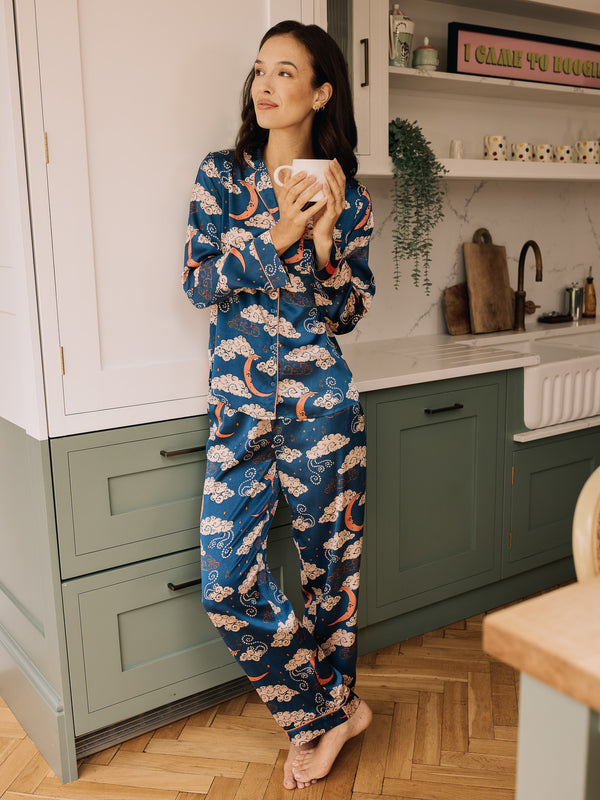 Woman in blue pyjama set with sky pattern standing in a kitchen holding a mug.