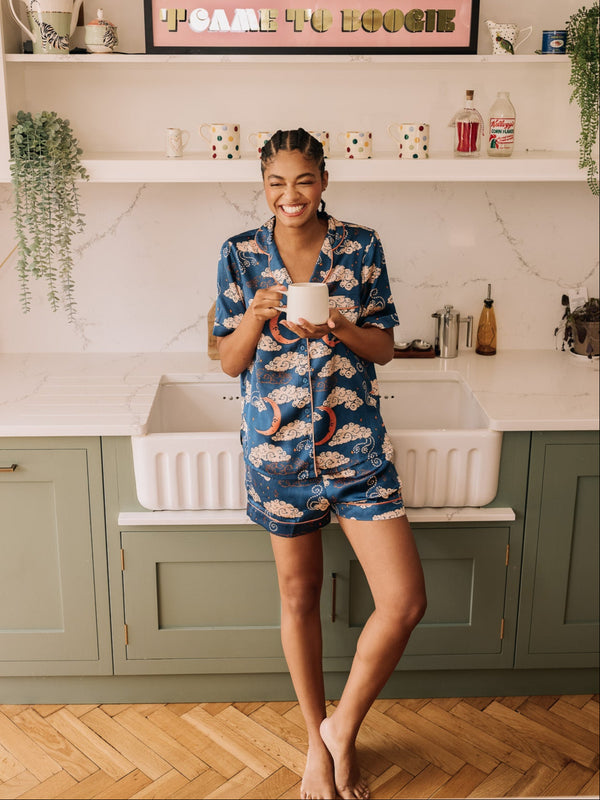 Woman in a kitchen wearing a blue pyjama set with cloud and moon pattern, holding a white mug.
