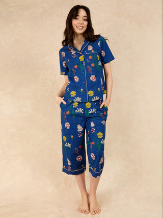 A woman standing and posing in a navy blue capri pyjama set with a field flower print. The pyjama set includes a button-down short-sleeved shirt and matching capri pants with a waist tie.