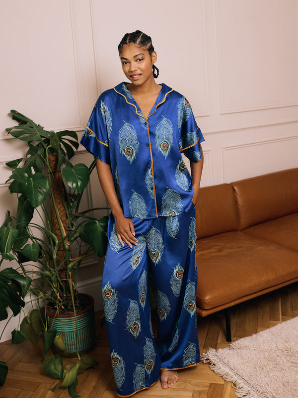 Woman wearing a blue satin pajama set with peacock feather pattern in a living room.