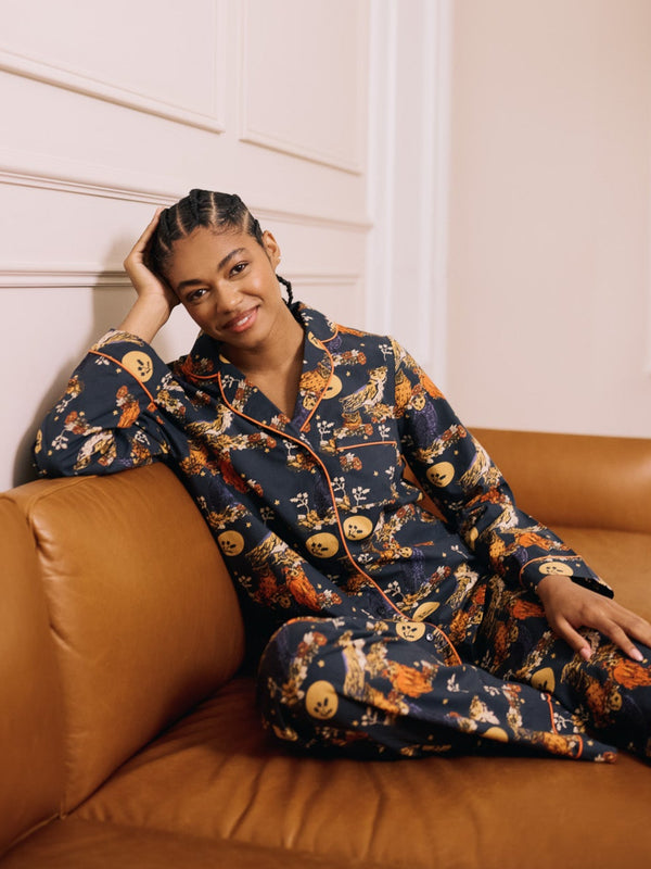 Woman in a grey cotton pyjama set with owl moonlight pattern sitting on a brown couch in a room with white walls and a plant.