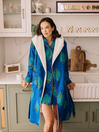Woman in a blue peacock-patterned cosy robe and matching nightshirt standing in a kitchen.