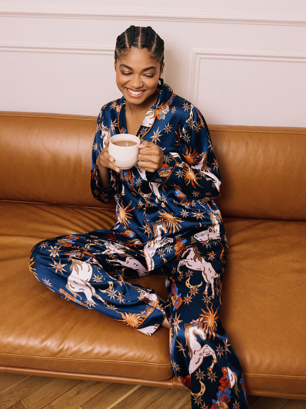 Woman in a blue unicorn patterned outfit pyjama set on a brown couch holding a mug.