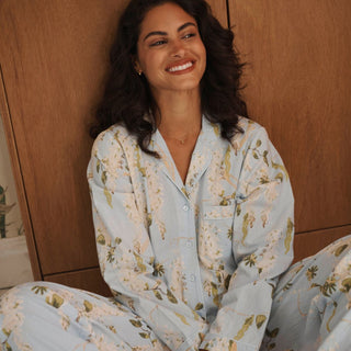 A woman smiling while wearing a light blue pajama set with a white floral print, sitting in front of a wooden background.