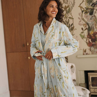 A woman wearing a light blue floral print cotton robe, standing in a cozy room with a wooden wardrobe and floral wallpaper.