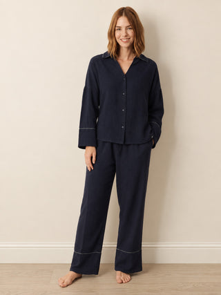 A woman modelling a matching navy blue pyjama set with contrasting light piping, consisting of a long-sleeve button-front top and wide-leg pants, standing against a neutral beige wall.