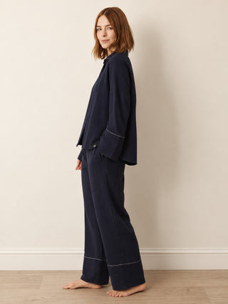 A woman stands barefoot in profile wearing a dark navy pyjama set with light piping details, consisting of a long-sleeve top and wide-leg pants, against a neutral beige wall and light wood floor.