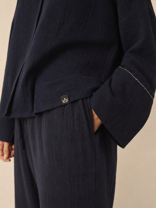 Close-up of a person wearing a dark navy textured two-piece pyjama set with a shirt and matching trousers, featuring a small embroidered crown logo on the pocket tab and contrast stitching on the sleeve.