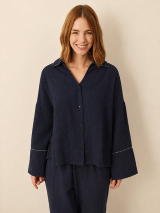 A woman with shoulder-length auburn hair wearing a dark navy blue button-up shirt and matching pyjama pants, standing against a plain beige studio background.