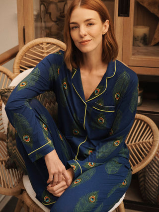 A woman wearing a navy blue jersey pyjama set with a green peacock feather print and gold piping, sitting in a wicker chair in a warm indoor setting.