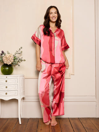 Woman wearing a pink and red abstract patterned satin pyjama set standing in a room with a white dresser and flowers.