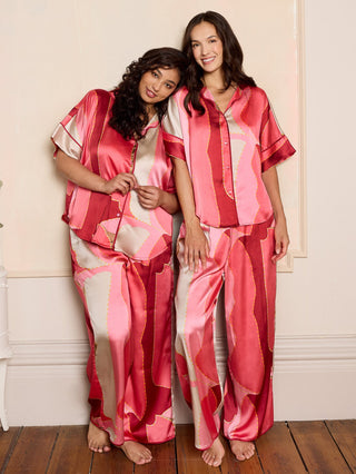 Two women wearing pink and red patterned pajama sets standing in a room with a white wall and wooden floor.