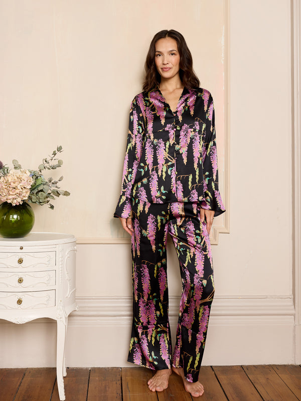 Woman wearing a floral black satin pyjama set in a room with a white dresser and flowers.
