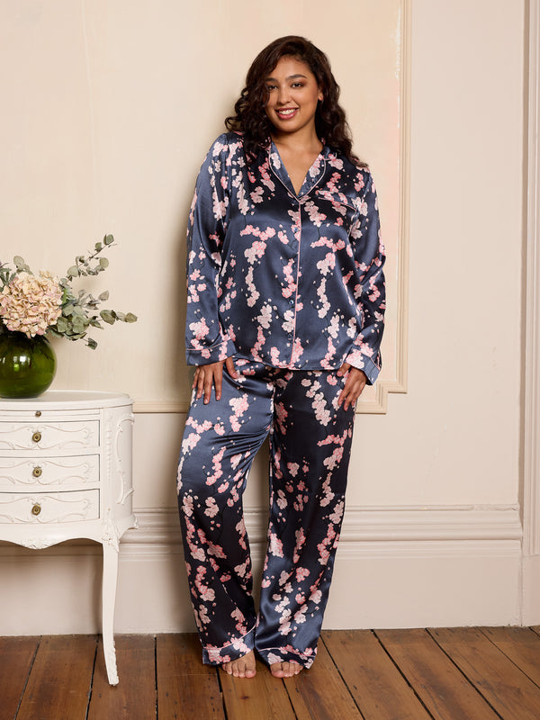 Woman wearing a satin floral pyjama set in a room with a white dresser and flowers.