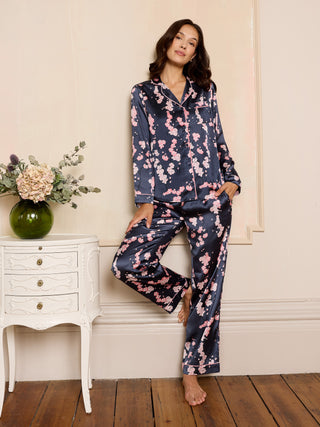Woman wearing a satin floral pyjama set in a cosy room with a white cabinet and flowers.