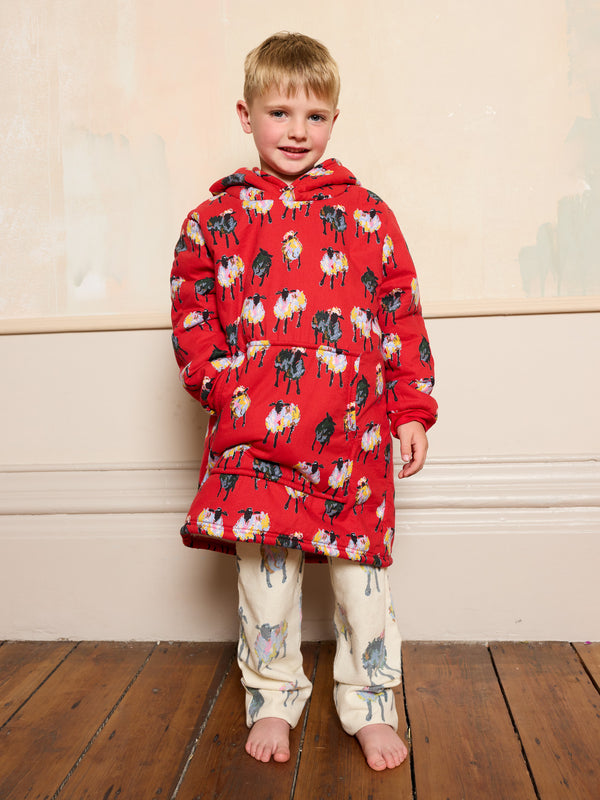Child wearing a red oversized hoodie with sheep pattern and white pyjamas on a wooden floor.
