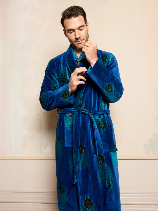 Man wearing a blue velvet robe with peacock feather patterns against a beige wall.