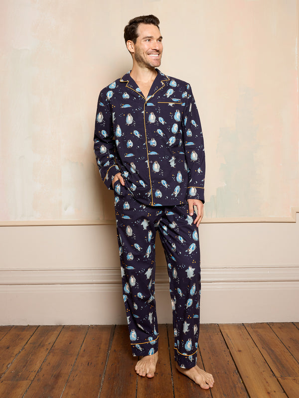 Man wearing a navy blue pyjama set with hand drawn christmas penguin print standing against a beige wall.
