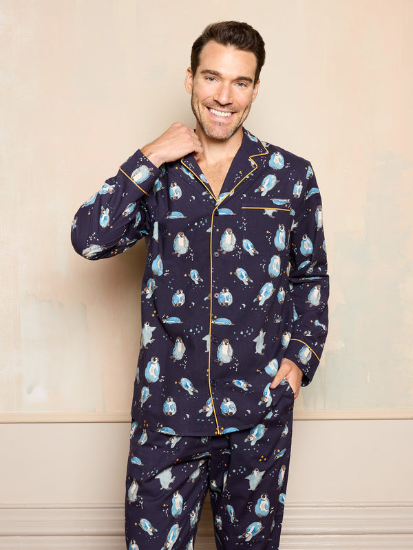 Man wearing a navy blue pyjama set with hand drawn christmas penguin print standing against a beige wall.