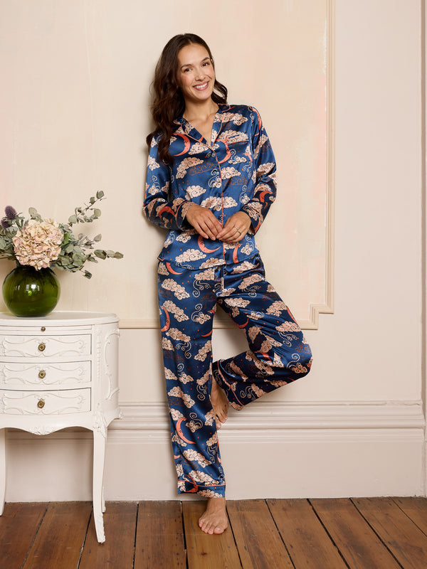 woman wearing navy long satin pyjama set with moon and clouds print in a room with a white dresser and vase of flowers