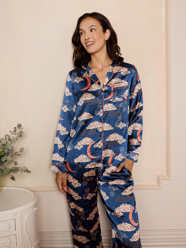 woman wearing navy long satin pyjama set with moon and clouds print with her hand in the trouser pocket.