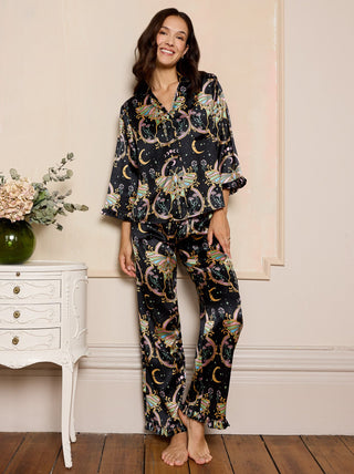 Woman wearing a mystical black satin  pyjama set standing in a room with a white side table and flowers.