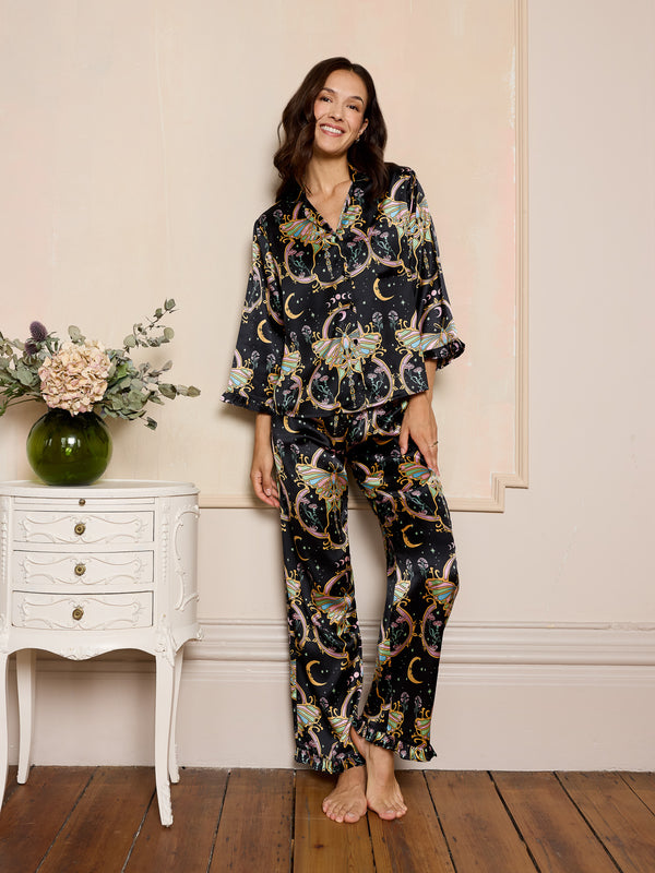 Woman wearing a mystical black satin  pyjama set standing in a room with a white side table and flowers.
