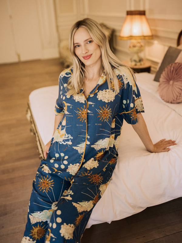 Woman wearing a blue celestial pyjama set sitting on a bed in a bedroom.