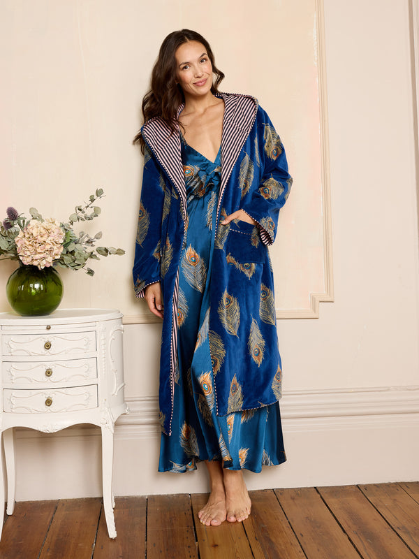 Woman wearing a blue patterned velvet robe standing in a bedroom with a white dresser and flowers.