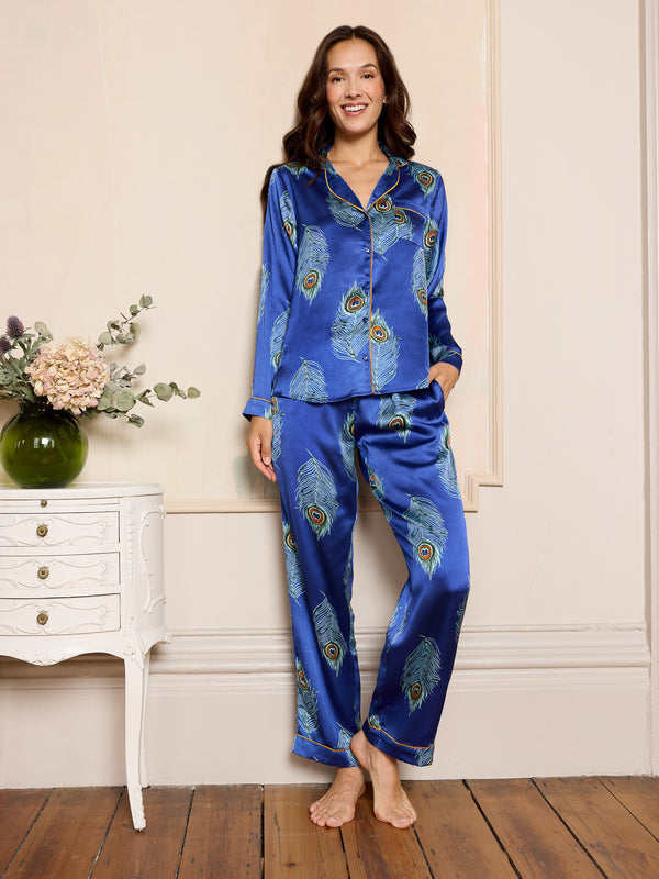 Woman wearing a navy blue satin pyjama set with peacock feather patterns in a home setting.