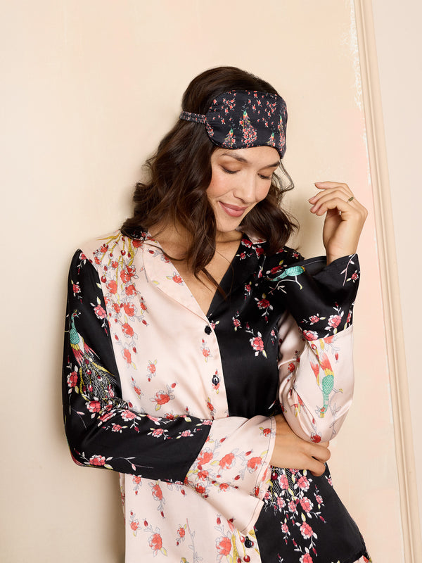 Woman wearing a pink and black floral satin pyjama set with peacock and rose pattern and matching eye mask