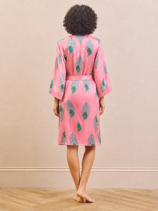 A woman viewed from behind wearing a pink robe with a green and blue peacock feather pattern, standing on a light wood floor against a beige wall.