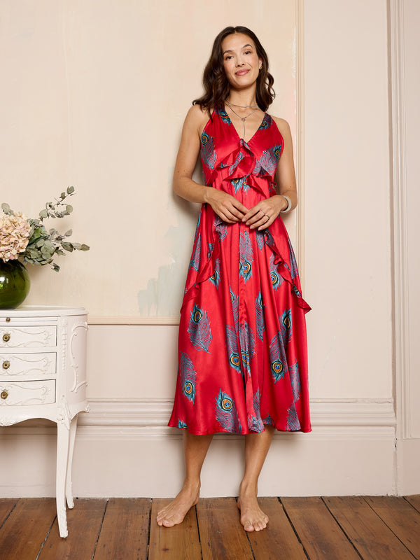 Woman wearing a red satin night dress with peacock feather patterns in a room with a white dresser and flowers.