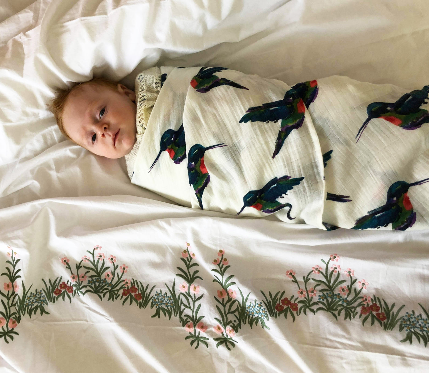Baby lies swaddled in cotton swaddle cloth, white base with hand painted multicoloured hummingbird pattern, birds in flight, in alternating directions