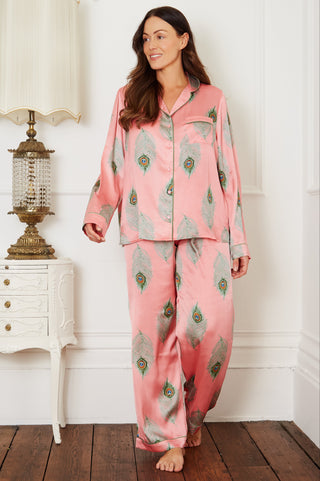 Brunette model wears warm pink based ladies traditional satin pyjamas, with repeating green peacock feather pattern, green piping on cuffs and collar