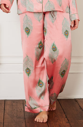 Brunette model wears warm pink based ladies traditional satin pyjamas, with repeating green peacock feather pattern, green piping on cuffs and collar
