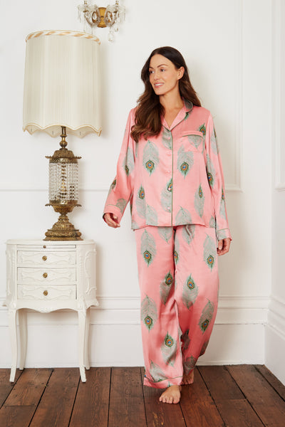 Brunette model wears warm pink based ladies traditional satin pyjamas, with repeating green peacock feather pattern, green piping on cuffs and collar