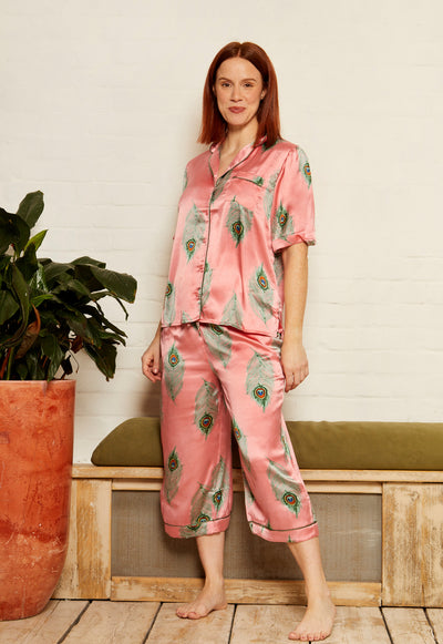 Auburn haired model stands in warm pink based ladies Capri satin pyjamas, Capri length, with repeating green peacock feather pattern, green piping on cuffs and collar